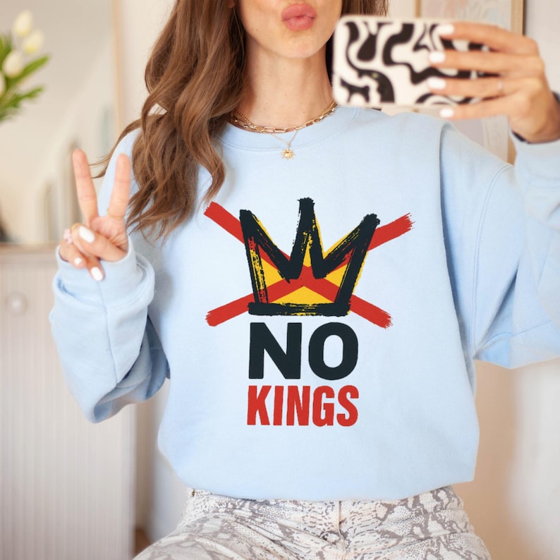 May include: A light blue sweatshirt with a graphic design featuring a crown crossed out with red lines, and the words "NO KINGS" in black and red. The sweatshirt is a casual style.