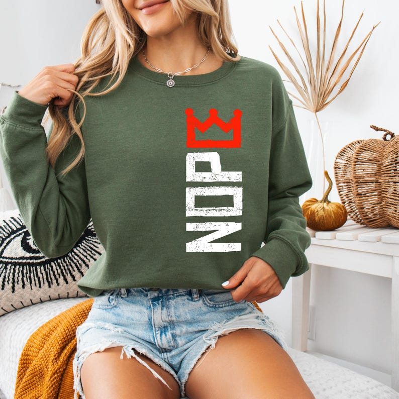 May include: Olive green sweatshirt with the letters "NDP" in white, topped with a red crown graphic. The long-sleeved top is made of a soft material. The sweatshirt is a casual style.