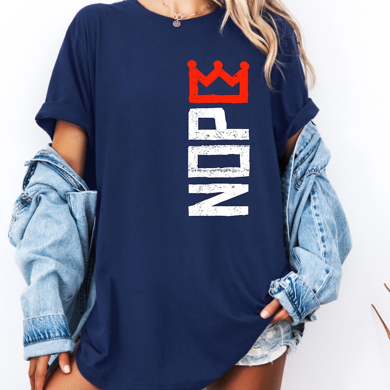 May include: Navy blue t-shirt with the word "NOPE" in white, topped with a red crown graphic. The shirt has a relaxed fit and short sleeves. A denim jacket is worn over the shirt.