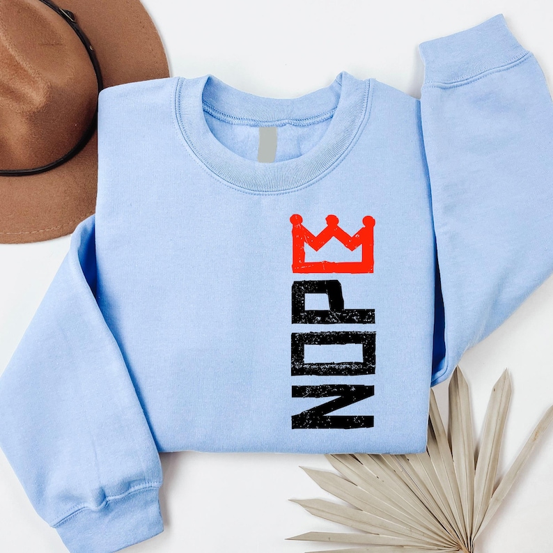 May include: A light blue sweatshirt with the word "NOPE" in black block letters and a red crown graphic. The sweatshirt is a crew neck style and has long sleeves. The design is printed vertically on the front.