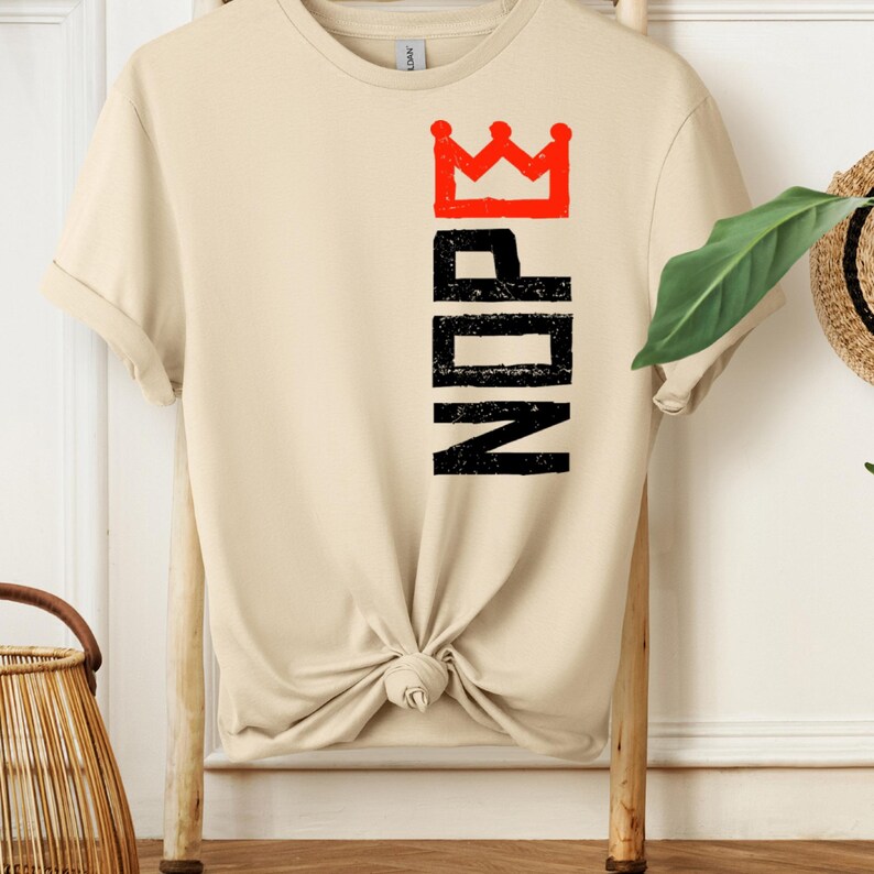 May include: A cream-colored t-shirt with the word "NOPE" printed vertically in black, topped with a red crown graphic. The shirt is knotted at the waist and displayed on a wooden hanger.