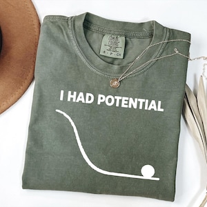 May include: An olive green t-shirt with the text "I HAD POTENTIAL" in white. A white graphic of a ball rolling down a slope is below the text. A silver necklace with a pendant is on the shirt.