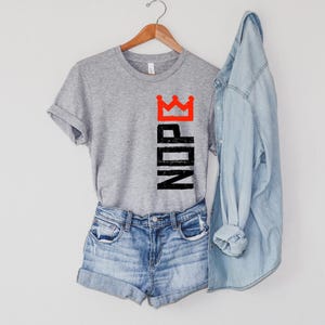 May include: A gray t-shirt with the word "NOPE" in black block letters and a red crown graphic. The shirt is paired with denim shorts and a light blue denim shirt. The items are hanging on a wooden hanger.