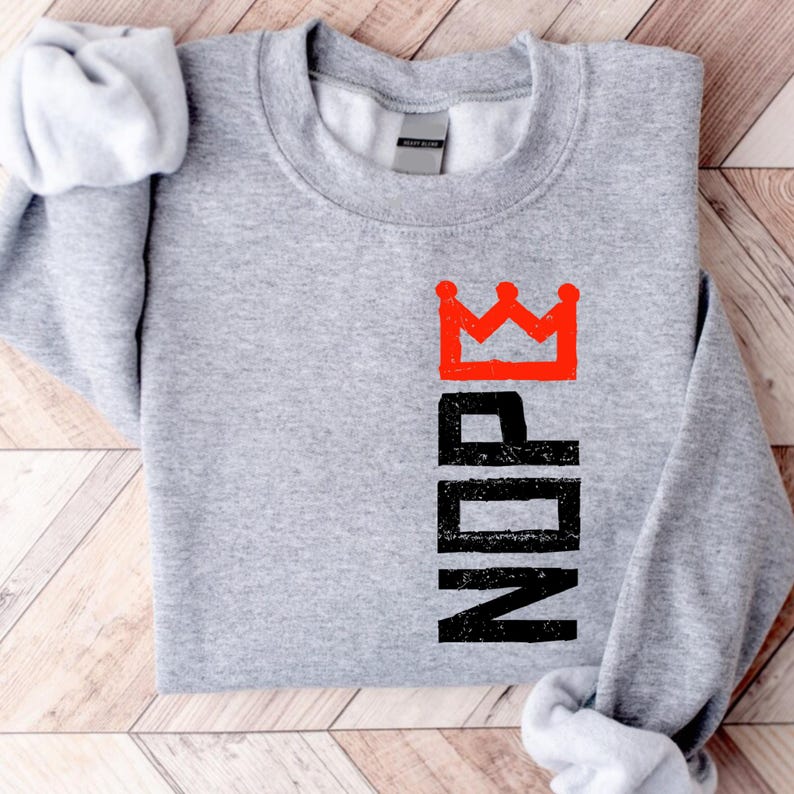 May include: A heather gray sweatshirt with the word "NOPE" printed vertically in black, topped with a red crown graphic. The long-sleeved shirt is folded, with the sleeves visible.