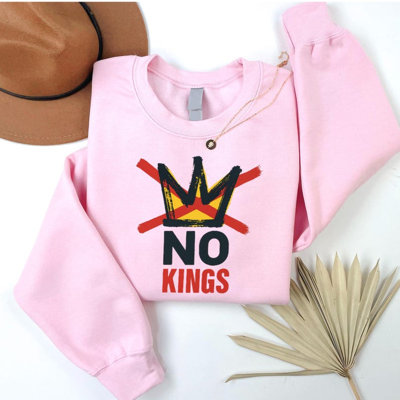 May include: A light pink sweatshirt with a graphic design featuring a crown crossed out with red lines, and the words "NO KINGS" in black and red. A gold necklace and a brown hat are visible.