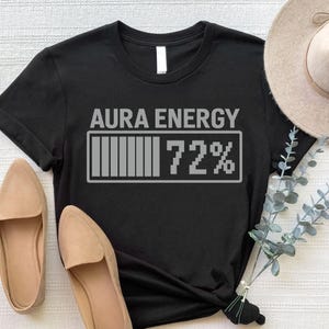 May include: Black t-shirt with the text "AURA ENERGY" above a battery-style graphic showing 72% charge. The shirt is paired with tan loafers and a beige hat. The design is in a simple, modern style.