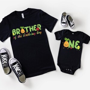 May include: Black t-shirts and a onesie with fruit graphics and text. The adult shirt says "Brother of the birthday boy" and the onesie says "ONE". Black and white sneakers are also in the image.