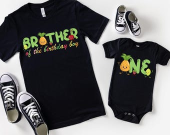 May include: Black t-shirts and a onesie with fruit graphics and text. The adult shirt says "Brother of the birthday boy" and the onesie says "ONE". Black and white sneakers are also in the image.