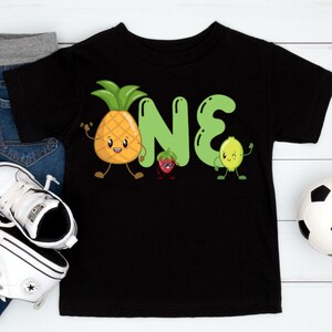 May include: Black toddler t-shirt with a playful "ONE" design featuring a cartoon pineapple, strawberry, and lime. The pineapple is yellow with a green top, the strawberry is red, and the lime is green. The shirt is paired with jeans, sneakers, and a soccer ball.