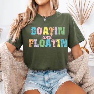 Puede incluir: Camiseta verde oliva con las palabras "BOATIN and FLOATIN" en colores pastel. El texto tiene un aspecto desgastado y vintage. La camiseta es de estilo casual y c&oacute;modo.