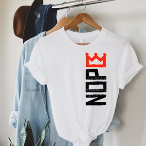 May include: White t-shirt with a red crown graphic above the black word "NOPE". The shirt is hanging on a wooden hanger with a denim shirt and a brown hat in the background.