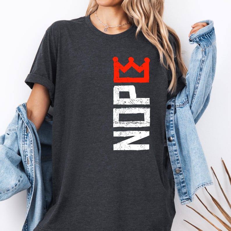 May include: Dark gray t-shirt with the word "NOPE" in white, and a red crown graphic. The shirt is a casual style, with short sleeves and a crew neck. The text and crown are printed vertically.