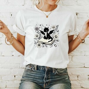 May include: White t-shirt featuring a black dragon illustration sitting on an open book, surrounded by daisies and foliage. The dragon has large wings and is centered on the shirt. The design is in black and white.