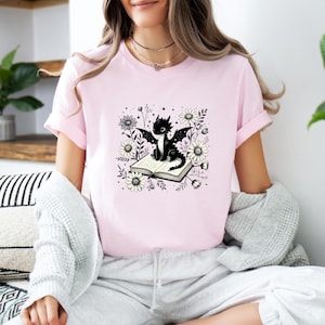 May include: A light pink t-shirt featuring a black dragon illustration. The dragon is sitting on an open book surrounded by white and gray flowers. The shirt has short sleeves and a crew neck.