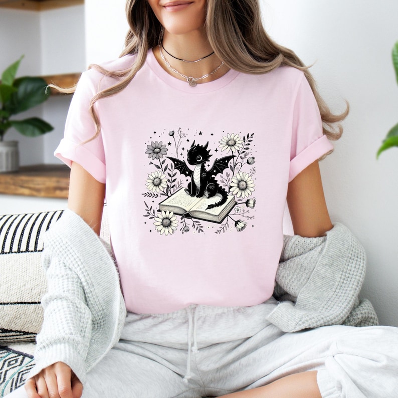 May include: A light pink t-shirt featuring a black dragon illustration. The dragon is sitting on an open book surrounded by white and gray flowers. The shirt has short sleeves and a crew neck.