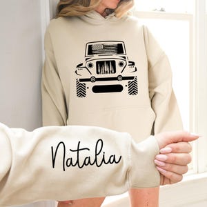 May include: A cream-colored hoodie with a black silhouette of a jeep with an American flag on the hood. The word "Natalia" is written in black cursive on the sleeve.