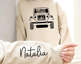 May include: A cream-colored hoodie with a black silhouette of a jeep with an American flag on the hood. The word "Natalia" is written in black cursive on the sleeve.