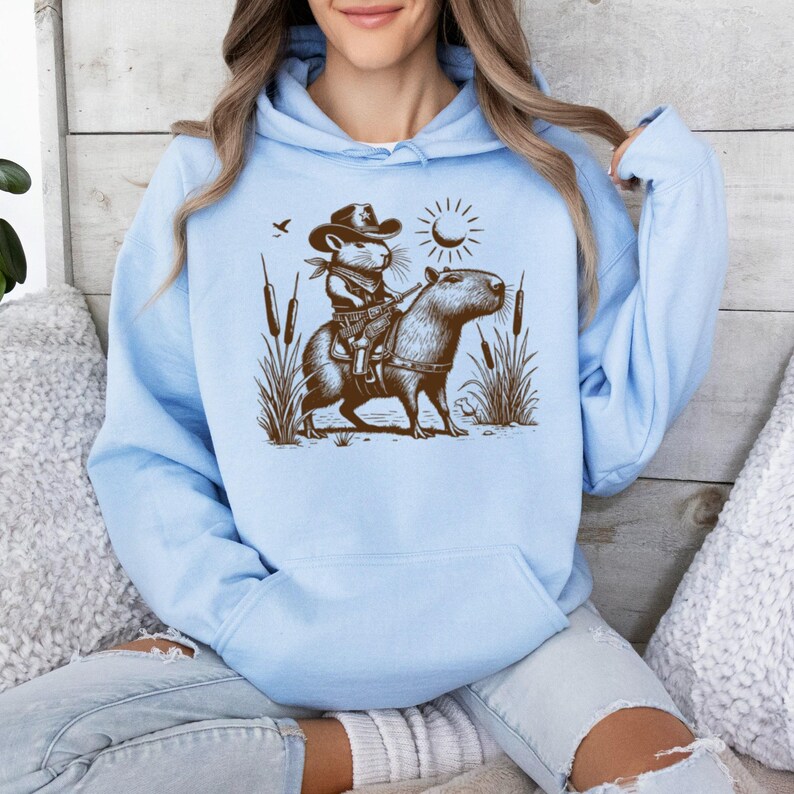 K&ouml;nnte beinhalten: Ein hellblauer Hoodie mit einer braunen Illustration einer Maus, die einen Cowboyhut tr&auml;gt und ein Capybara reitet. Die Illustration enth&auml;lt eine Sonne, einen Halbmond und Rohrkolben.