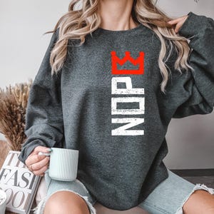 May include: Dark gray sweatshirt with the word "NDP" in white vertical text and a red crown graphic above. The sweatshirt is a crew neck style.