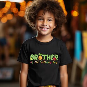May include: A black t-shirt with the text "BROTHER of the birthday boy" in green, yellow, and red. The word "BROTHER" has a fruit design. A young boy with curly hair is wearing the shirt and smiling.