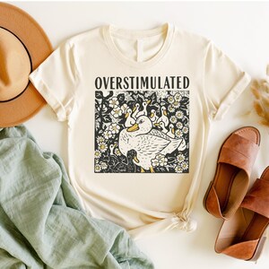 K&ouml;nnte beinhalten: Cremefarbenes T-Shirt mit dem Wort "OVERSTIMULATED" &uuml;ber einer schwarz-wei&szlig;en Grafik einer Ente, umgeben von Entenk&uuml;ken und Blumen. Das Design hat gelbe Akzente. Das Shirt wird mit Hut, Sandalen und einem gr&uuml;nen Tuch pr&auml;sentiert.