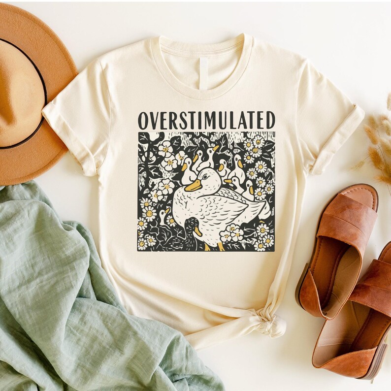 K&ouml;nnte beinhalten: Cremefarbenes T-Shirt mit dem Wort "OVERSTIMULATED" &uuml;ber einer schwarz-wei&szlig;en Grafik einer Ente, umgeben von Entenk&uuml;ken und Blumen. Das Design hat gelbe Akzente. Das Shirt wird mit Hut, Sandalen und einem gr&uuml;nen Tuch pr&auml;sentiert.