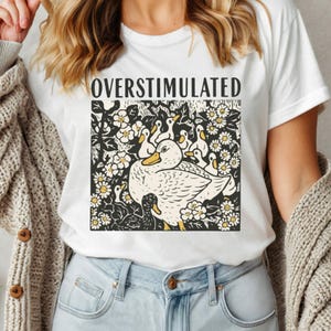 K&ouml;nnte beinhalten: Wei&szlig;es T-Shirt mit dem Wort "OVERSTIMULATED" &uuml;ber einer Grafik einer Ente und Entenk&uuml;ken. Die Ente und die Entenk&uuml;ken sind wei&szlig; mit gelben Schn&auml;beln und F&uuml;&szlig;en, umgeben von wei&szlig;en Blumen mit gelben Zentren.