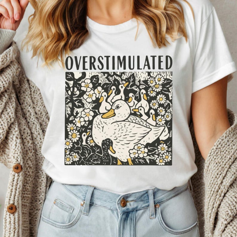 K&ouml;nnte beinhalten: Wei&szlig;es T-Shirt mit dem Wort "OVERSTIMULATED" &uuml;ber einer Grafik einer Ente und Entenk&uuml;ken. Die Ente und die Entenk&uuml;ken sind wei&szlig; mit gelben Schn&auml;beln und F&uuml;&szlig;en, umgeben von wei&szlig;en Blumen mit gelben Zentren.
