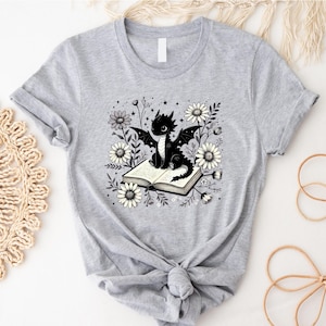 May include: A light gray t-shirt featuring a black dragon sitting on an open book, surrounded by white daisies and foliage. The dragon has large wings and is centered on the shirt. The design is a whimsical illustration.