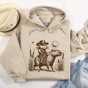 K&ouml;nnte beinhalten: Ein beiger Kapuzenpullover mit einem braunen Grafikdruck eines Cartoon-Nagetiers, das einen Cowboyhut tr&auml;gt und ein Capybara reitet. Das Nagetier h&auml;lt eine Pistole und das Capybara steht in hohem Gras.