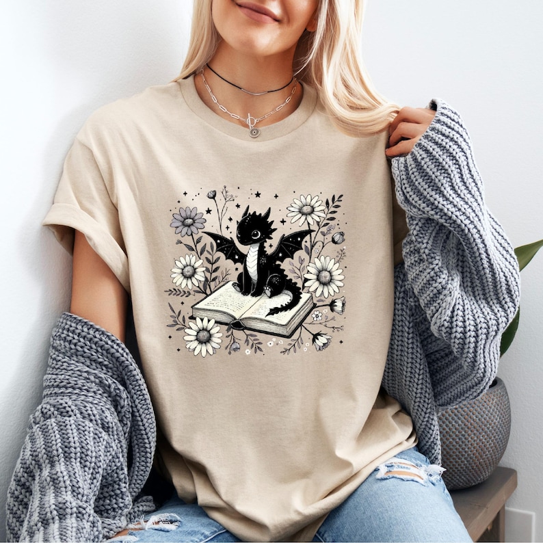 May include: Beige t-shirt featuring a black dragon illustration sitting on an open book, surrounded by daisies and other flowers. The dragon has large wings and is set against a floral background. The shirt is a neutral color.