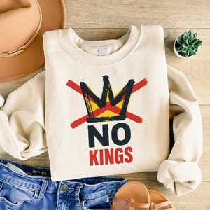 May include: Cream-colored sweatshirt with the text "NO KINGS" in black and red, and a crossed-out crown graphic. The crown is yellow and black with red lines through it. The sweatshirt is on a wooden surface.