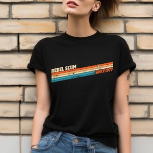 May include: Black t-shirt with the text "REBEL SCUM" above a retro design of orange, yellow, and blue stripes, with "SINCE 1977" printed to the right. The shirt is a classic crew neck style.