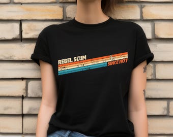 May include: Black t-shirt with the text "REBEL SCUM" above a retro design of orange, yellow, and blue stripes, with "SINCE 1977" printed to the right. The shirt is a classic crew neck style.