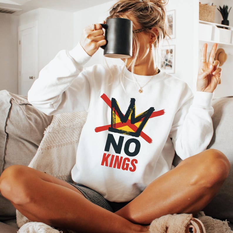 May include: White sweatshirt with a graphic design featuring a crown crossed out with red lines, the words "NO KINGS" in black and red. A black mug is held up near the face. The person is sitting on a couch.