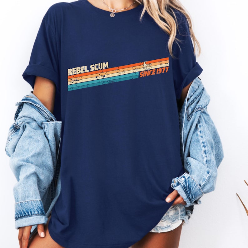 May include: Navy blue t-shirt with the text "REBEL SCUM" above a retro-style design of horizontal stripes in orange, yellow, teal, and blue. The text "SINCE 1977" is printed to the right of the stripes. The shirt is worn with a denim jacket.