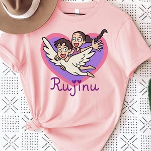 May include: Pink t-shirt featuring a cartoon illustration of two people flying with angel wings inside a purple heart. The word "Rujinu" is written in a cursive font below the heart.