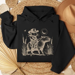 K&ouml;nnte beinhalten: Ein schwarzer Hoodie mit einem grafischen Aufdruck eines Cartoon-Nagetiers, das einen Cowboyhut tr&auml;gt und ein Capybara reitet. Das Nagetier h&auml;lt eine Waffe und das Capybara steht in hohem Gras.