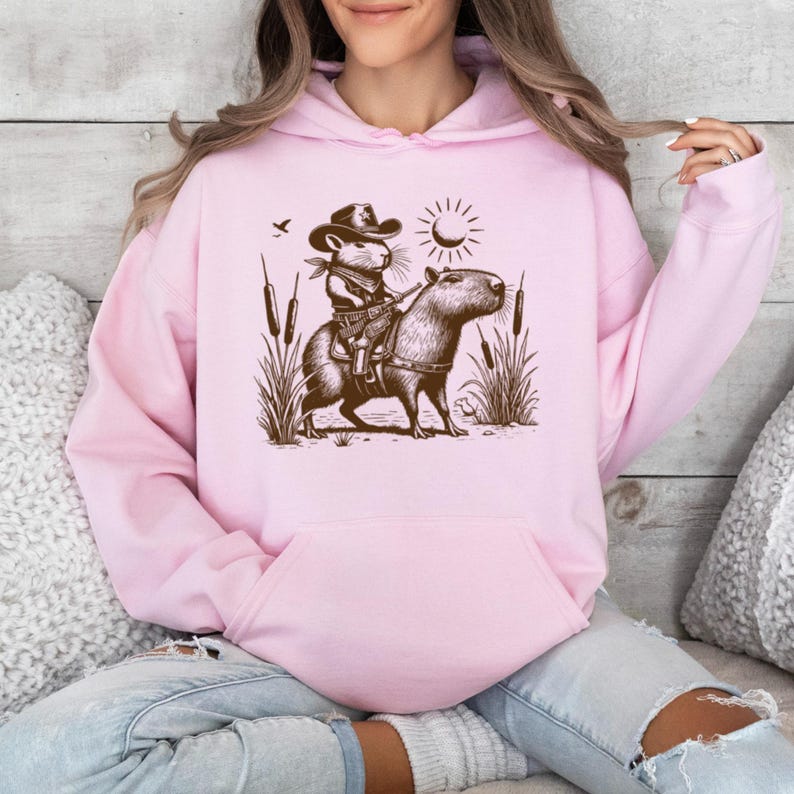 K&ouml;nnte beinhalten: Ein rosa Hoodie mit einem Aufdruck eines Mause mit einem Cowboyhut, der ein Capybara reitet. Der Aufdruck ist in Braun und Schwarz gehalten und zeigt eine Sonne und Schilf im Hintergrund.
