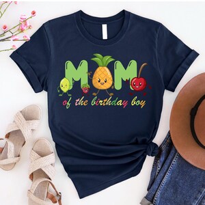 May include: Navy blue t-shirt with a playful design featuring the word "MOM" in green, a smiling pineapple, lemon, strawberry, and cherry. The text "of the birthday boy" is written below in colorful letters. A casual, fun shirt.