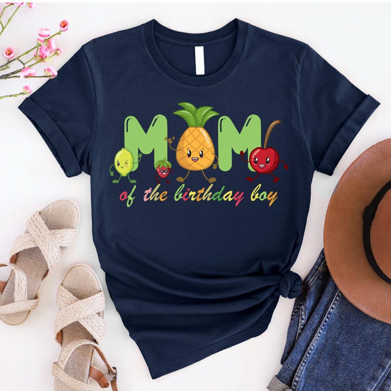 May include: Navy blue t-shirt with a playful design featuring the word "MOM" in green, a smiling pineapple, lemon, strawberry, and cherry. The text "of the birthday boy" is written below in colorful letters. A casual, fun shirt.