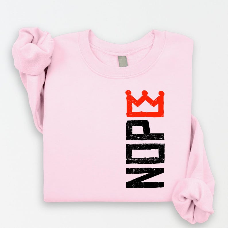 May include: A light pink sweatshirt with the word "NOPE" printed vertically in black, topped with a red crown graphic. The sleeves are rolled up, and the sweatshirt is made of a soft material.