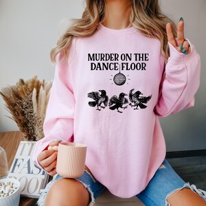 May include: A light pink sweatshirt with the text "MURDER ON THE DANCE FLOOR" above a disco ball graphic and three black crow illustrations. The sweatshirt is a crew neck style.