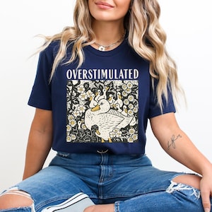 K&ouml;nnte beinhalten: Marineblaues T-Shirt mit dem Wort "OVERSTIMULATED" &uuml;ber einer Grafik einer Ente, die von Entenk&uuml;ken und Blumen umgeben ist. Die Grafik ist in Schwarz, Wei&szlig; und Gelb gehalten.
