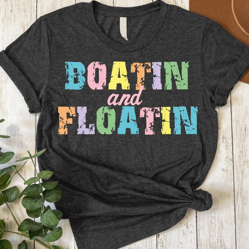 Puede incluir: Camiseta gris oscuro con las palabras "BOATIN" y "FLOATIN" en letras de bloque coloridas y desgastadas. La palabra "and" est&aacute; en una fuente cursiva m&aacute;s peque&ntilde;a. La camiseta est&aacute; anudada en la parte inferior y tiene mangas enrolladas.