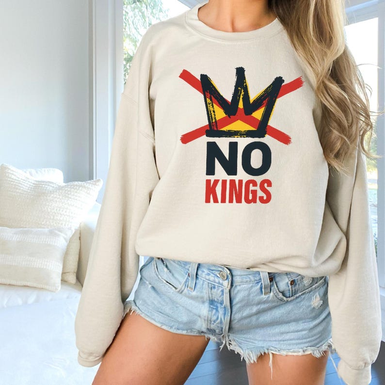 May include: A beige sweatshirt with a graphic design featuring a crown crossed out with red lines, and the words "NO KINGS" in black and red. The sweatshirt is paired with denim shorts.