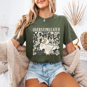 K&ouml;nnte beinhalten: Olivgr&uuml;nes T-Shirt mit dem Wort "OVERSTIMULATED" &uuml;ber einer Grafik einer Ente und Entenk&uuml;ken, umgeben von Blumen. Das Shirt ist kurz&auml;rmlig und hat einen l&auml;ssigen Stil.