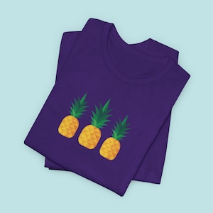 Puede incluir: Una camiseta morada con un gráfico de tres piñas amarillas en la parte delantera.