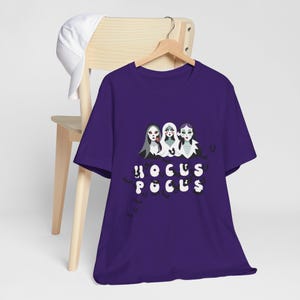 Pode incluir: Camiseta roxa com um gráfico de três bruxas do filme Hocus Pocus. As bruxas usam túnicas pretas e têm pele verde. O texto "Hocus Pocus" está escrito em letras pretas abaixo das bruxas.