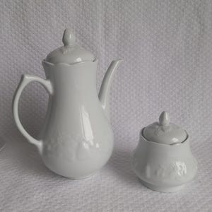 Peut inclure: Service à café en porcelaine blanche comprenant une cafetière et un sucrier. La cafetière a une anse incurvée et un long bec verseur. Le sucrier a un couvercle avec un petit bouton. Les deux pièces ont un motif floral en relief.